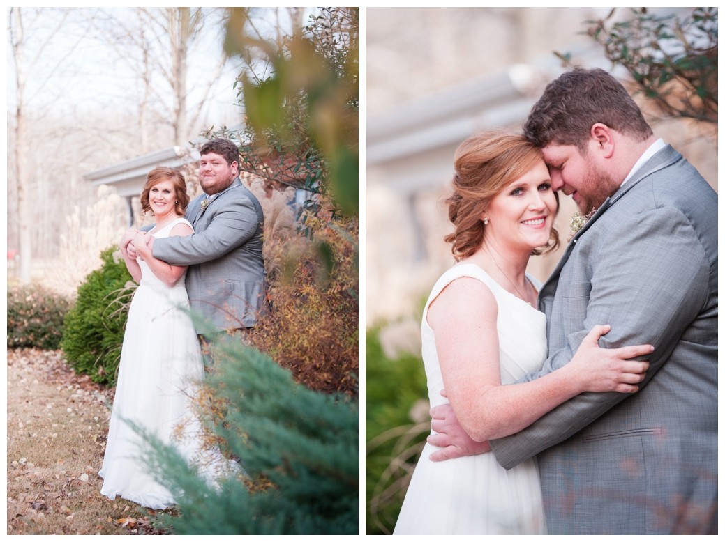 bride and groom standing near bushes with his arms around her and bride smiling at camera while groom nuzzles her