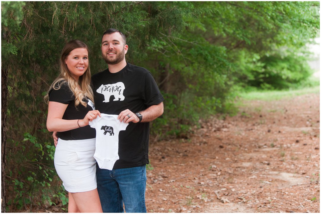 baby announcement couple holding onesie by trees